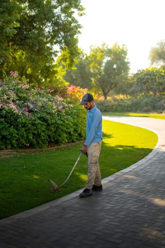 Man trimming lawn in a sunny Dubai park, surrounded by lush greenery and flowers.